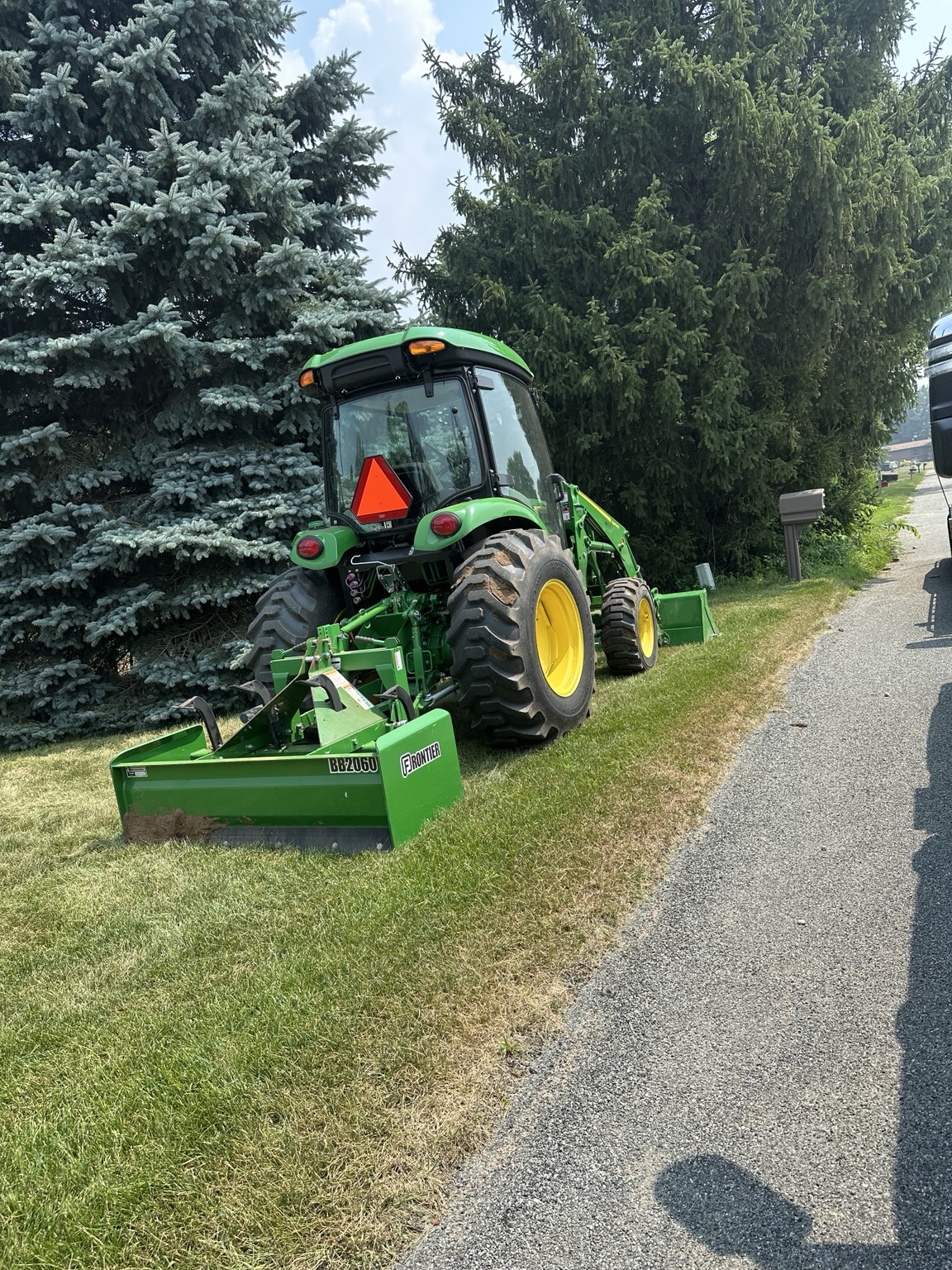 John Deere tractor with rear box blade beside large evergreen trees